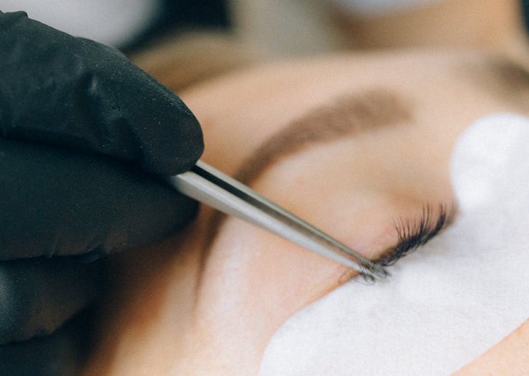 Black-gloved technician applying eyelash extensions to a woman's eye in a beauty salon setting.